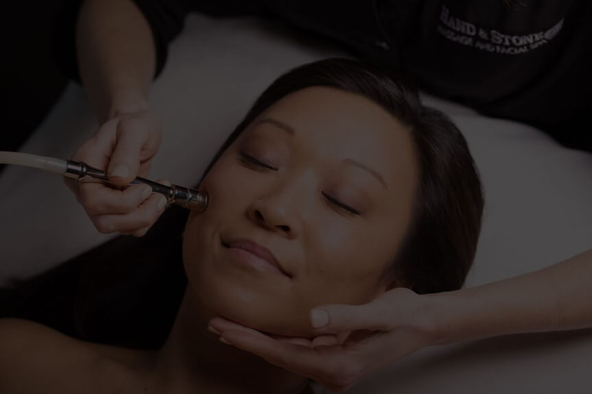 Person lying down with eyes closed, receiving a facial treatment with a microdermabrasion tool.